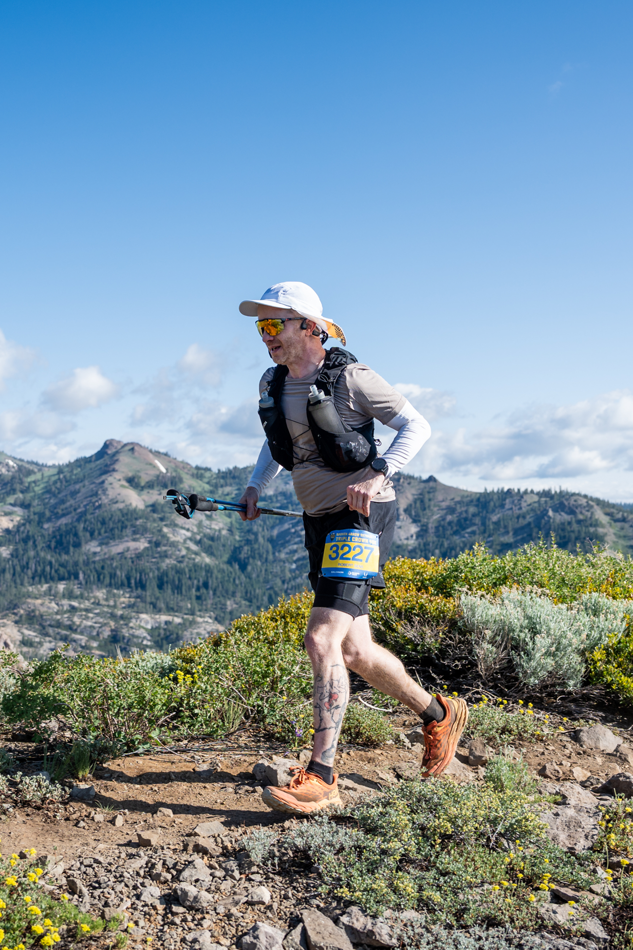 Runner with race bib 3227 and trekking poles running on rocky trail in mountainous terrain.