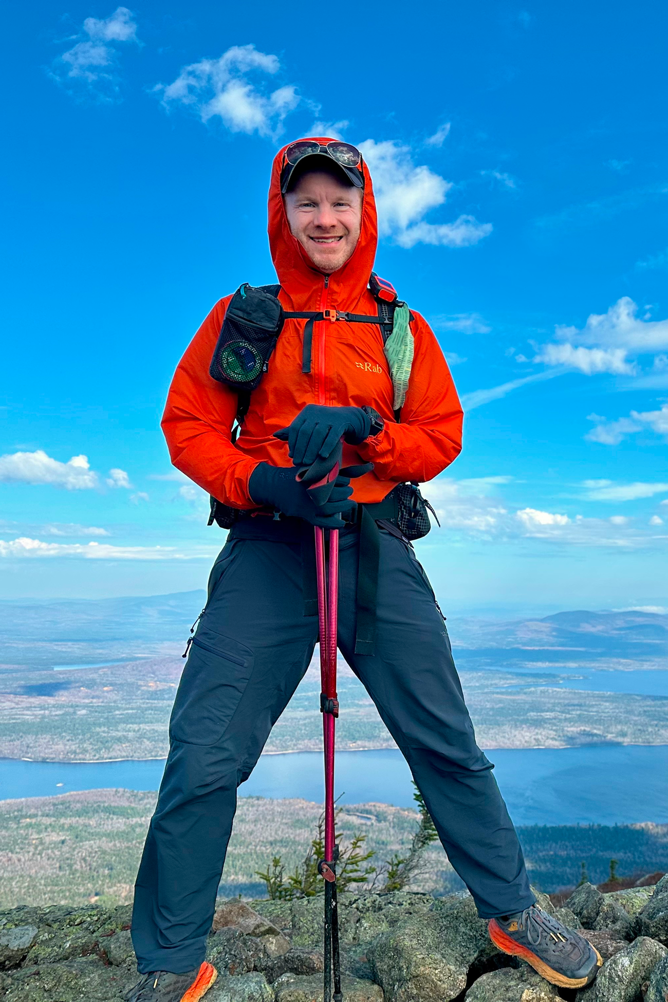 Smiling hiker in red jacket and gloves standing on rocky terrain with trekking poles and mountain lake view.