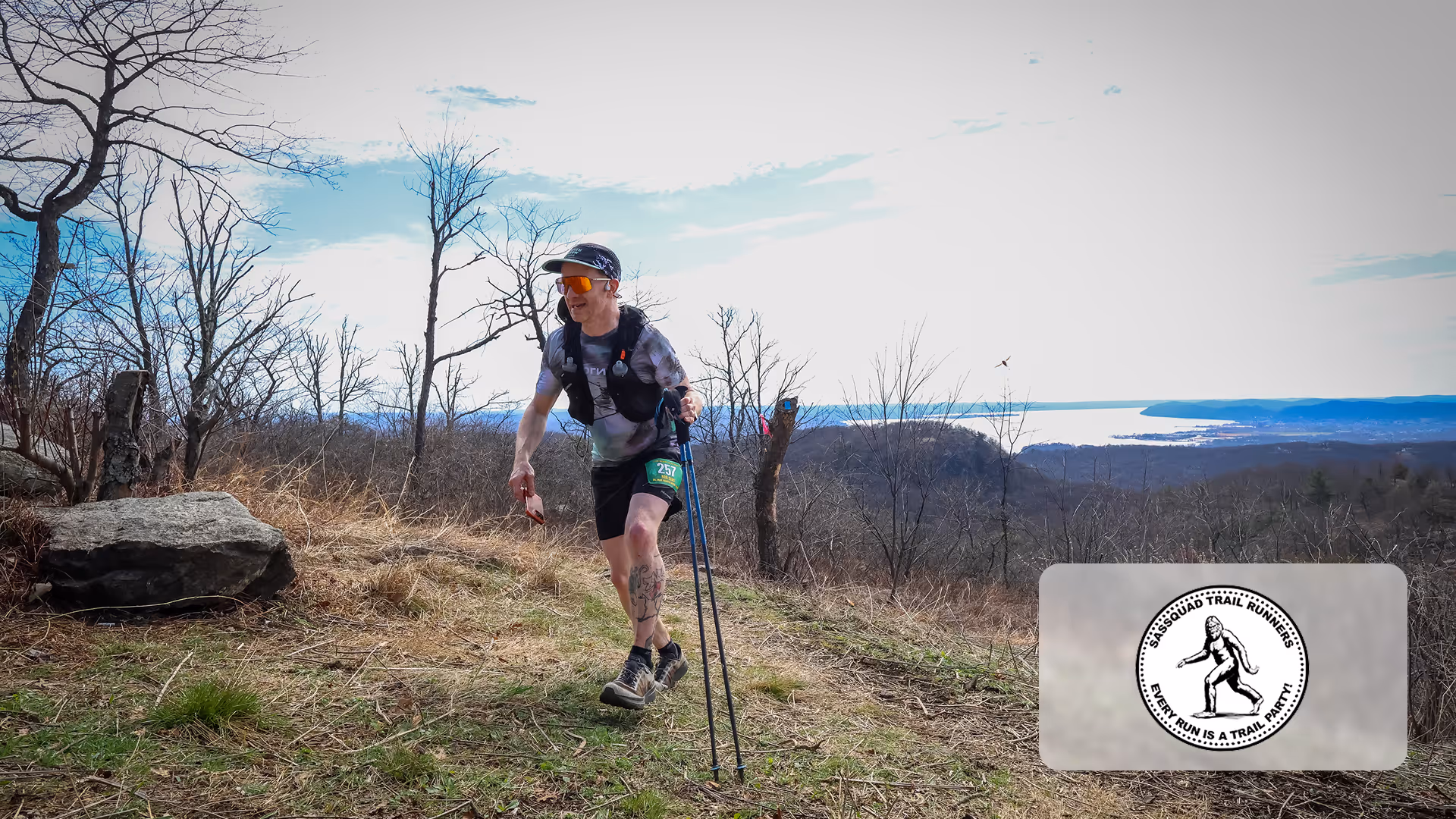 Trail runner with poles running on a grassy hill with leafless trees and water in the distance.