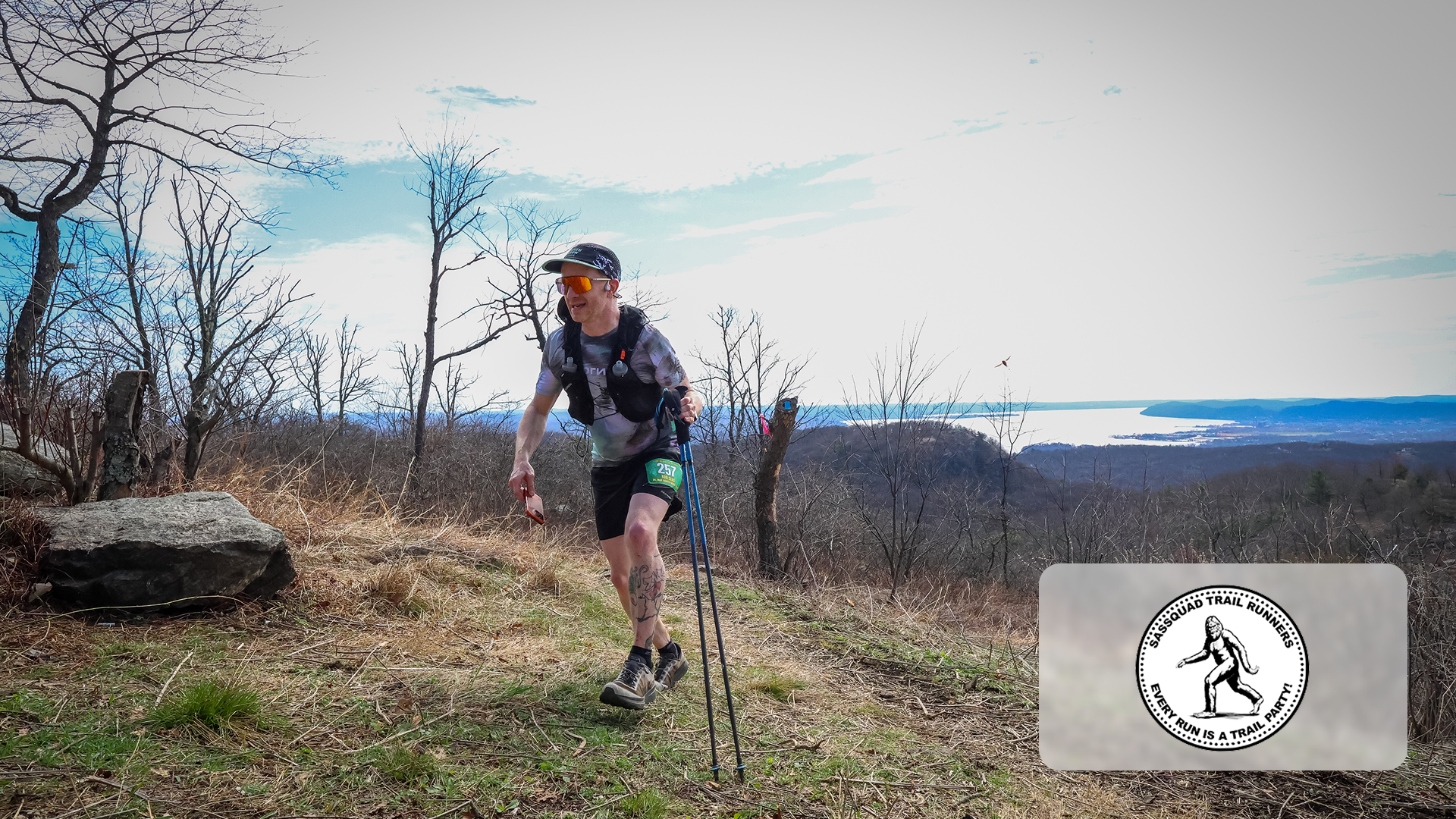 Trail runner with poles running on a grassy hill with leafless trees and water in the distance.