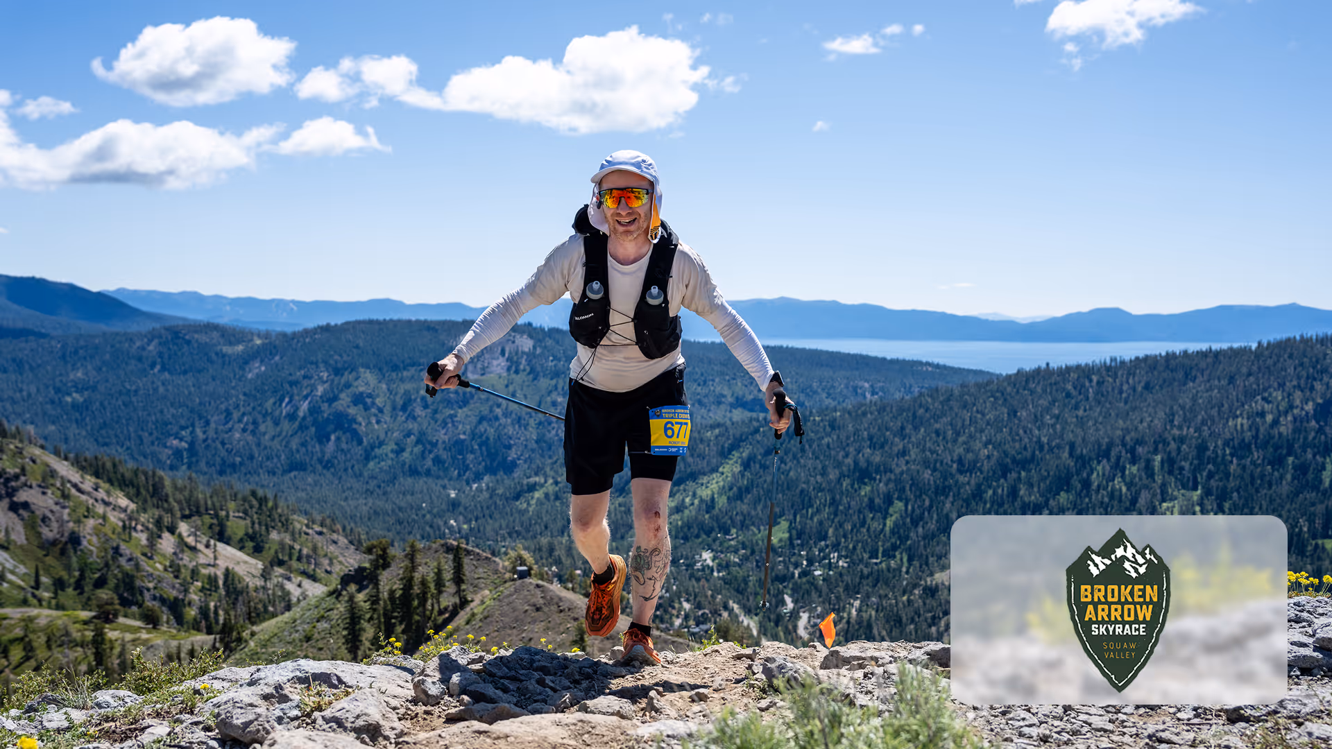 Runner with trekking poles wearing bib 677 on rocky mountain trail with forest and lake views.