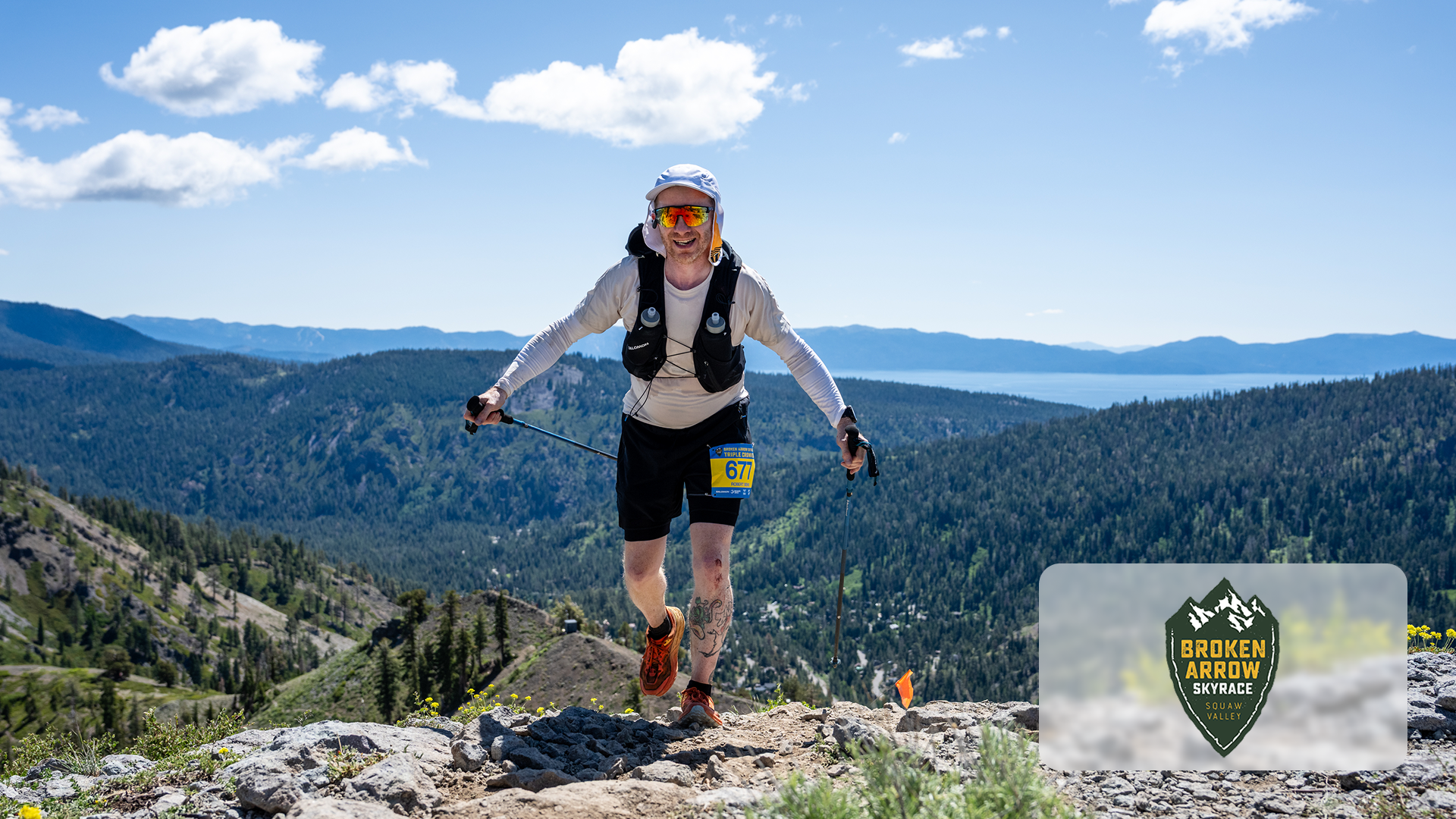 Runner with trekking poles wearing bib 677 on rocky mountain trail with forest and lake views.