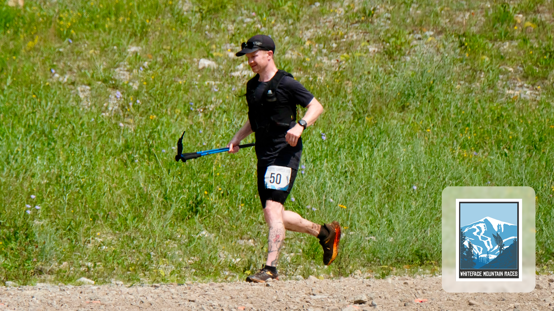 Runner with race number 50 runs on rocky trail holding a trekking pole in a grassy area.