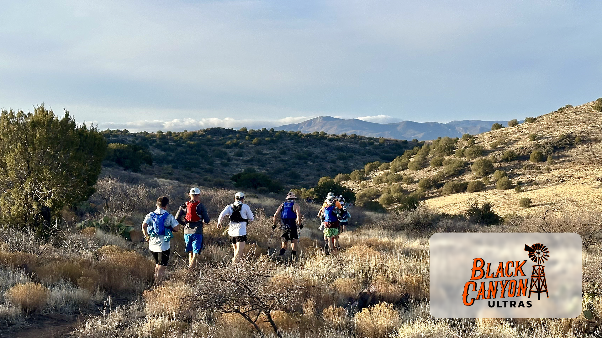 Group of runners hiking on a trail through dry brush with mountains in the distance.