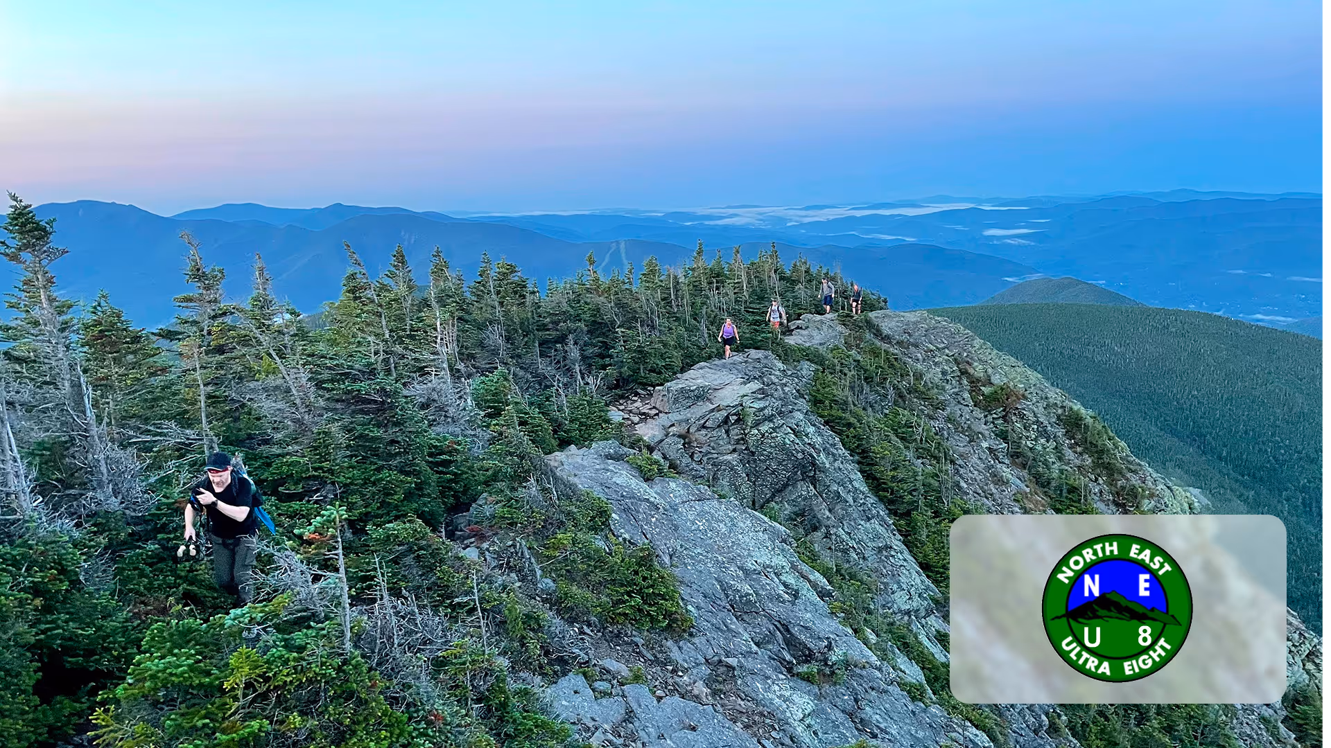 Hikers trekking on a rocky mountain ridge surrounded by pine trees and distant mountain layers at dusk.