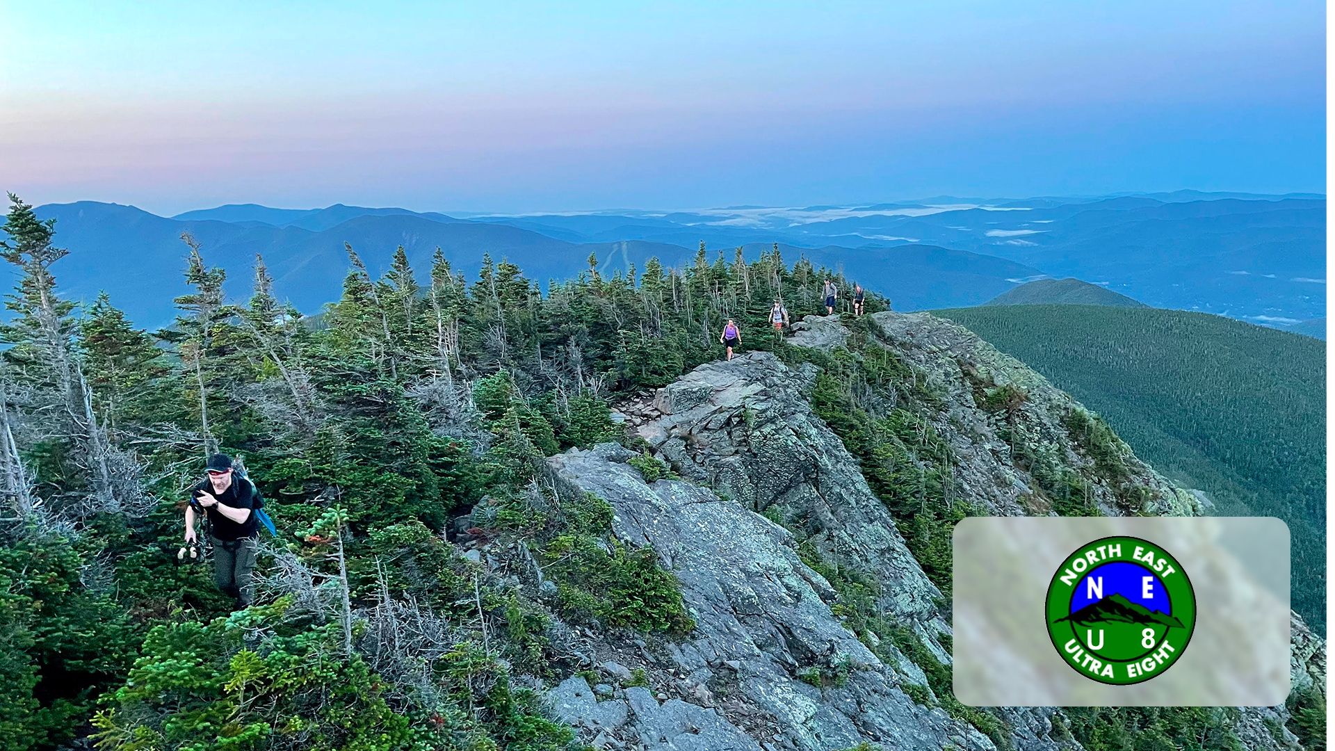 Hikers trekking on a rocky mountain ridge surrounded by pine trees and distant mountain layers at dusk.