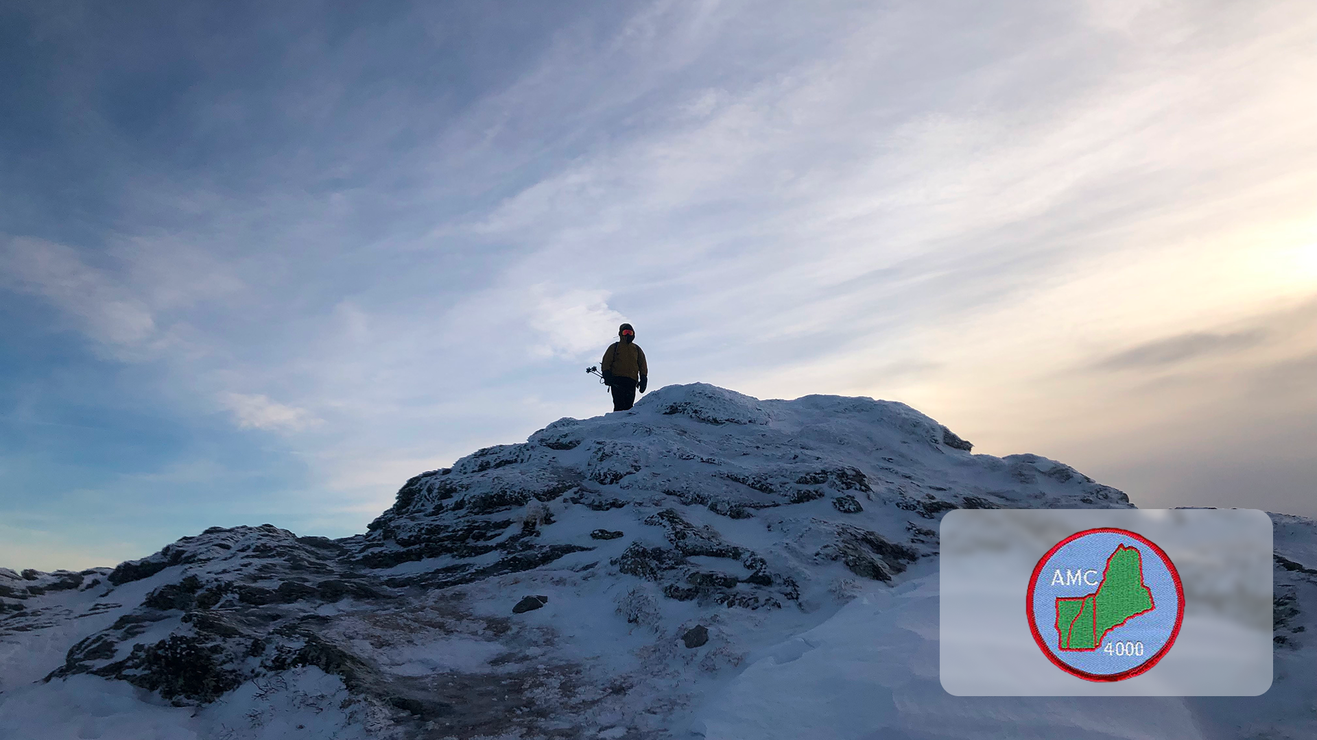 Person in winter gear standing on snowy rocky peak under a cloudy sky at sunset.