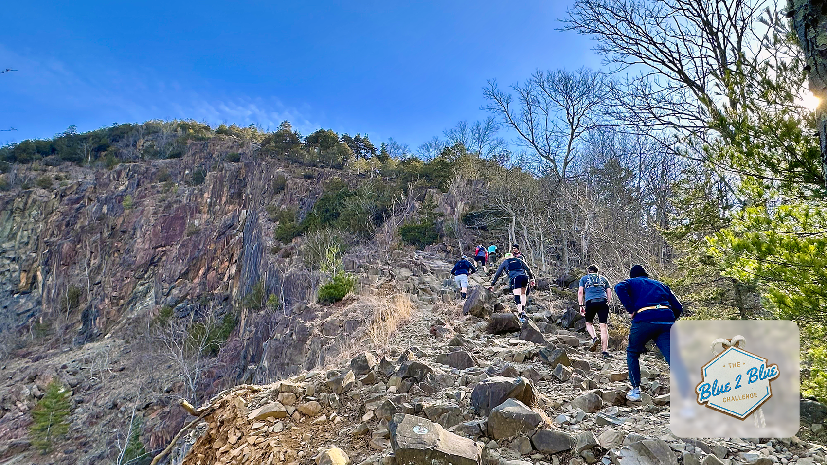 Hikers climbing a rocky trail uphill against a blue sky with sparse trees on a hillside.
