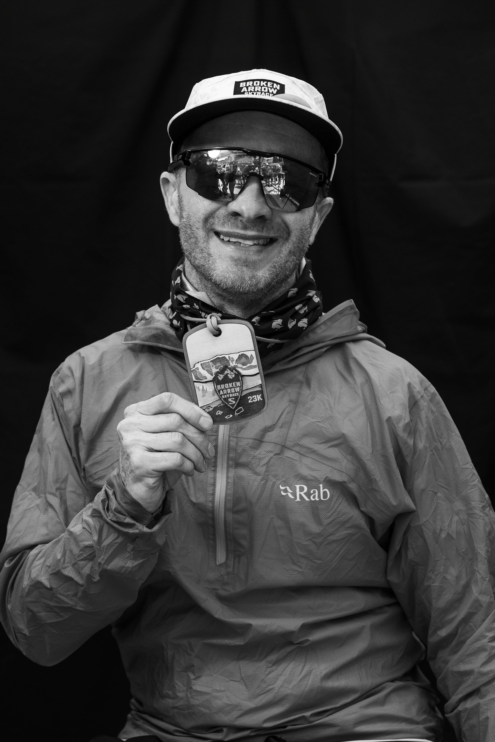 Smiling man in cap and sunglasses holding Broken Arrow Skyrace 23K medal, wearing a Rab jacket.