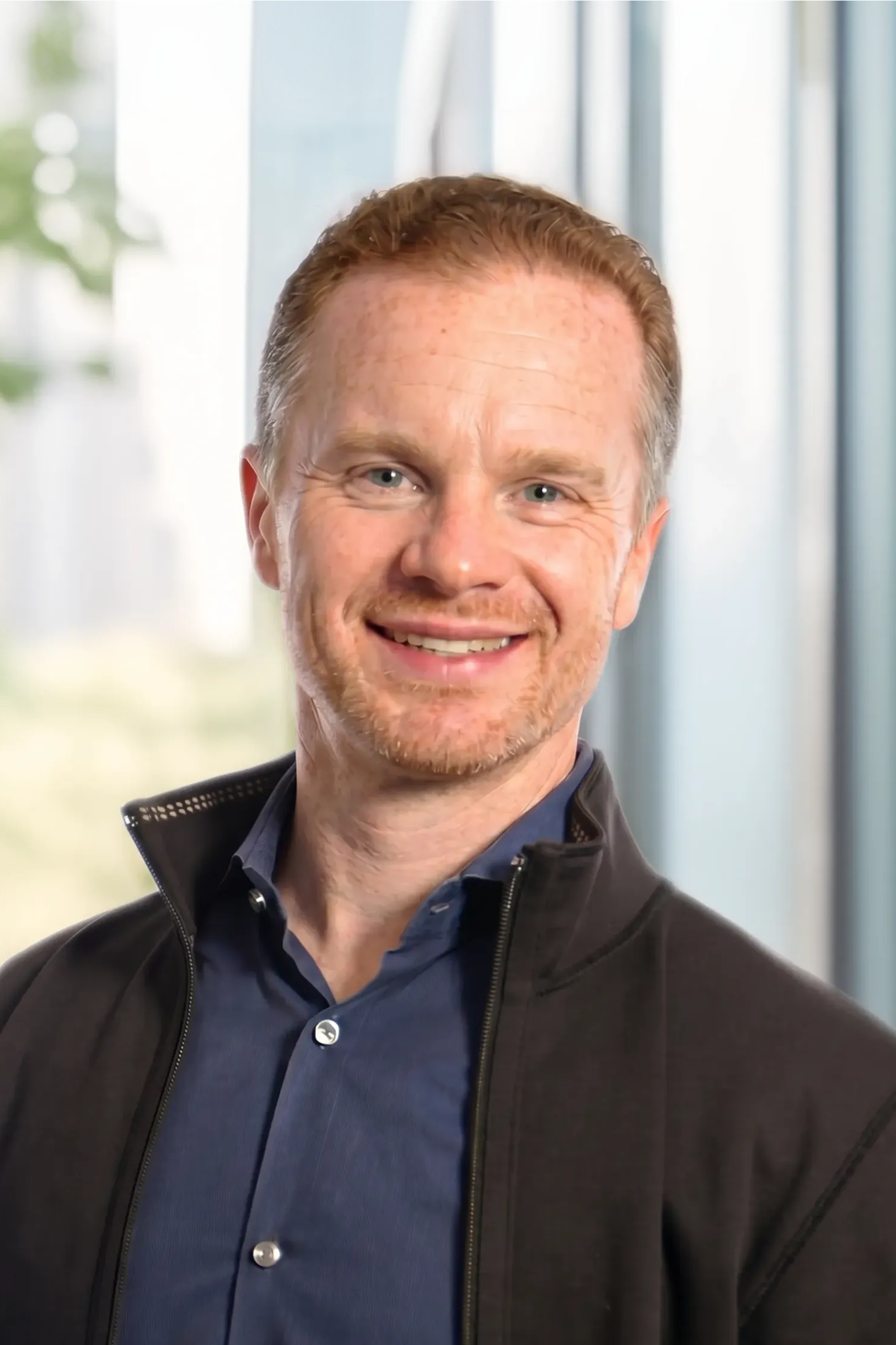 Smiling man with light brown hair wearing a blue shirt and black jacket, indoors.