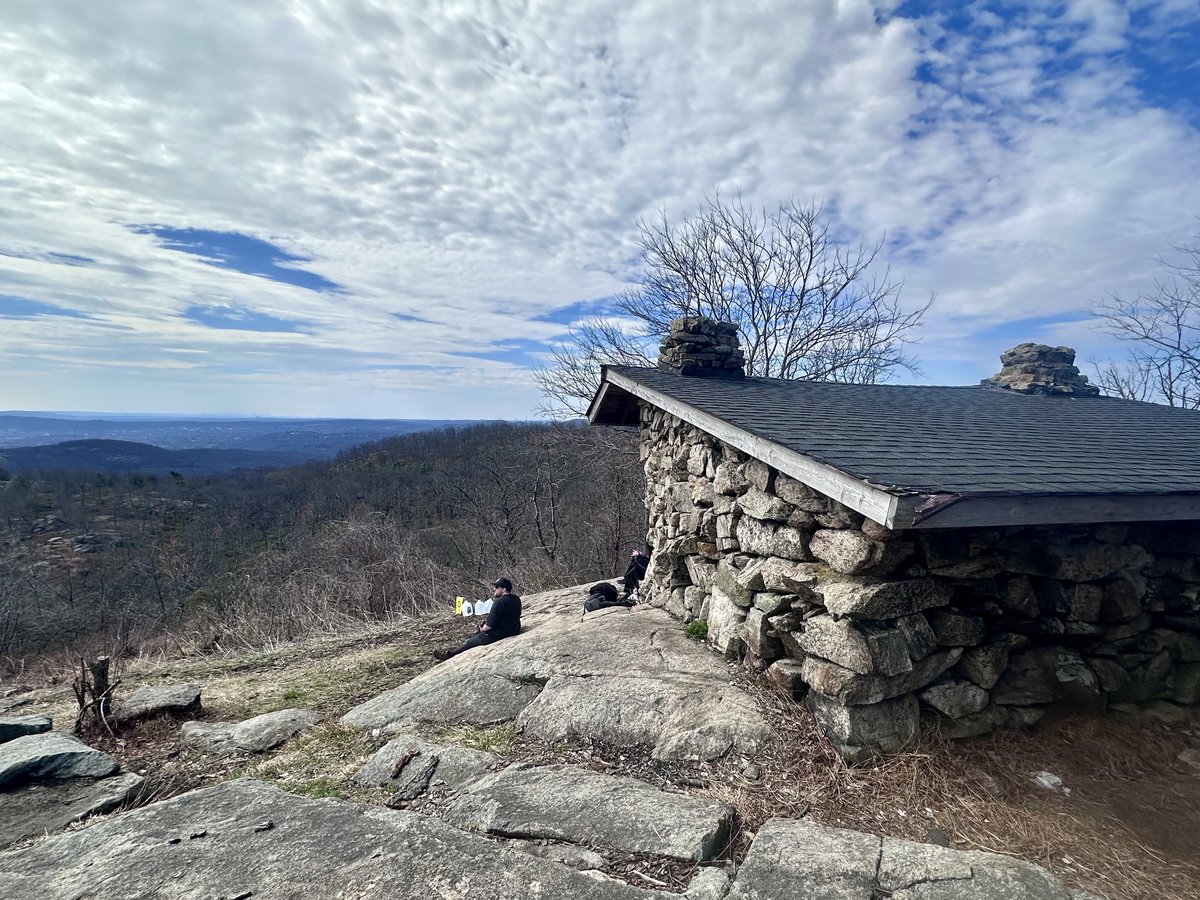 The stone West Mountain Shelter at the high point of the course, a hiker sitting on the open rock ledge with a broad forested view beyond