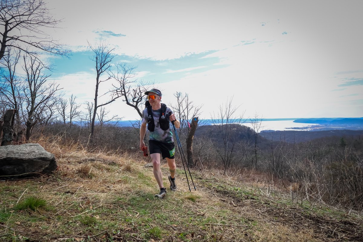 Rob Sens pushing up an exposed grassy ridgeline with trekking poles, Hudson River and distant hills behind him