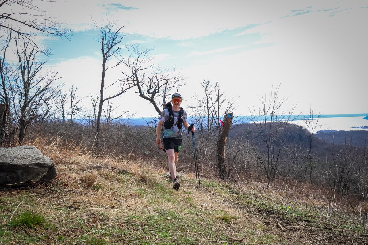 Rob Sens hiking with trekking poles on an open ridgeline, Hudson River visible in the background, smiling at the camera