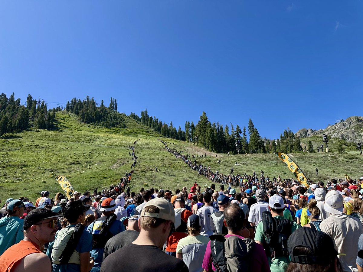Hundreds of runners and spectators pack the grassy lower slopes of Palisades Tahoe at the start of the Ascent, with a long ant-line of athletes winding up the mountain above them under a cloudless blue sky