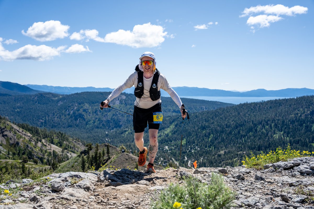 Running hard down a rocky ridgeline toward the camera, bib 677, poles out for balance, with Lake Tahoe and the surrounding forests clearly visible in the distance under a blue sky