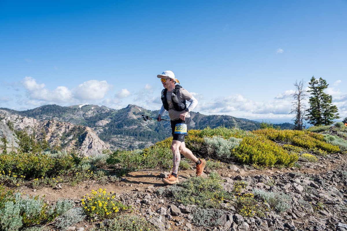 Running along a rocky high-alpine ridgeline with poles, bib 3227, with the Sierra Nevada peaks and a patchwork of wildflowers visible against a mostly blue sky