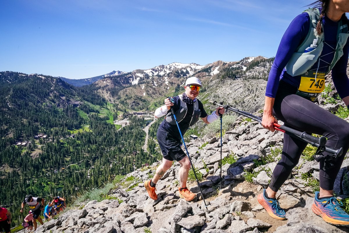 Smiling in a white bucket hat and orange-lensed sunglasses, poles in hand, picking my way up a steep rocky slope with the valley floor of Palisades Tahoe visible far below and snow-patched peaks in the background
