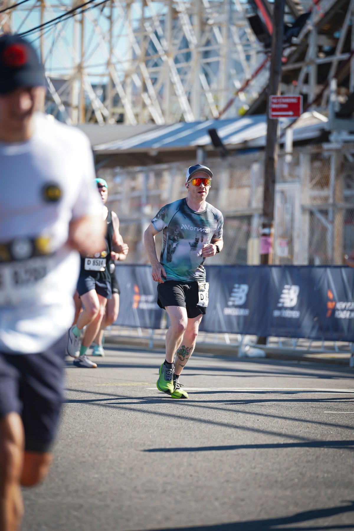 Runner in a grey Nnormal shirt and orange-lens sunglasses running along a road near Coney Island, wooden roller coaster structure in the background