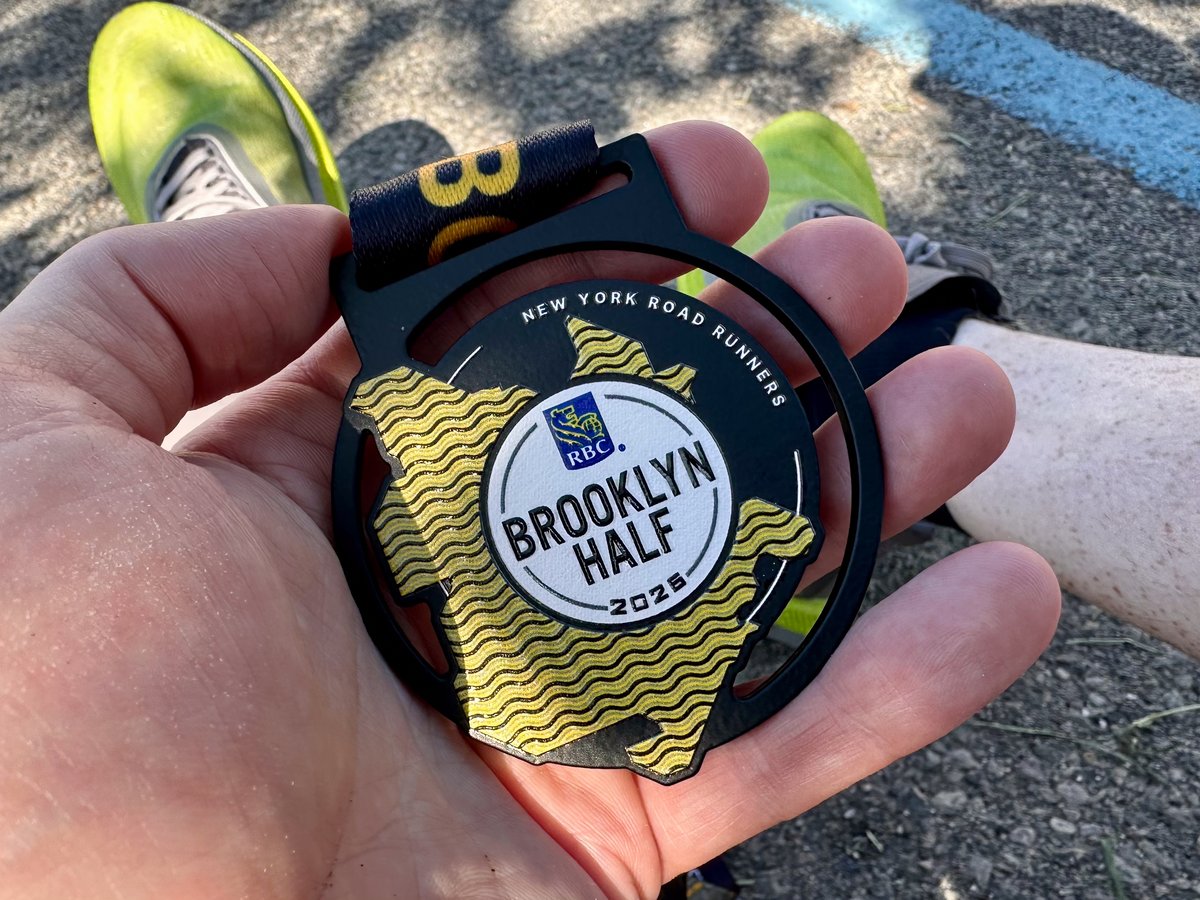 The 2026 RBC Brooklyn Half finisher medal — black metal with gold wave pattern, RBC and Brooklyn Half 2026 text at center — held up against asphalt and a bright green shoe sole visible in the background