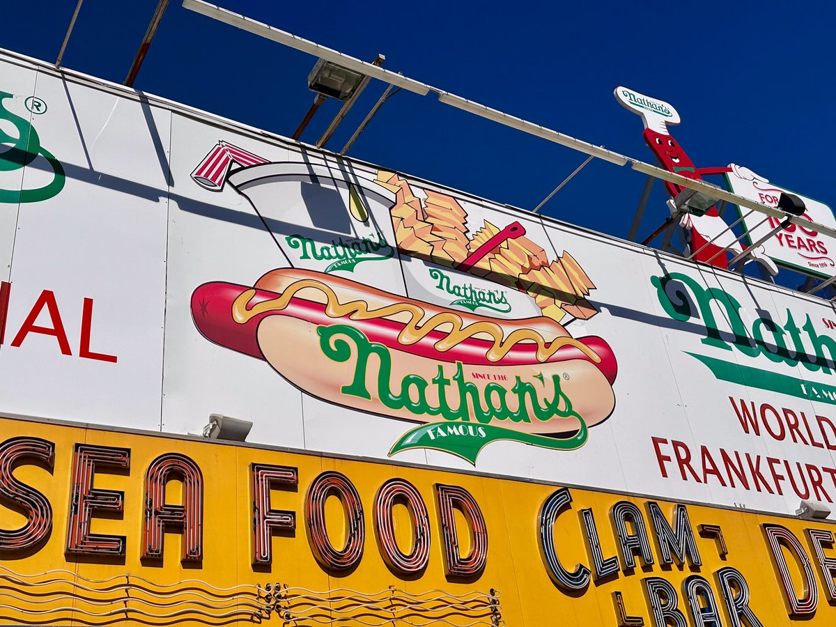 Looking up at the Nathan's Famous sign at Coney Island — bold hot dog graphic against a yellow and white facade, deep blue sky above