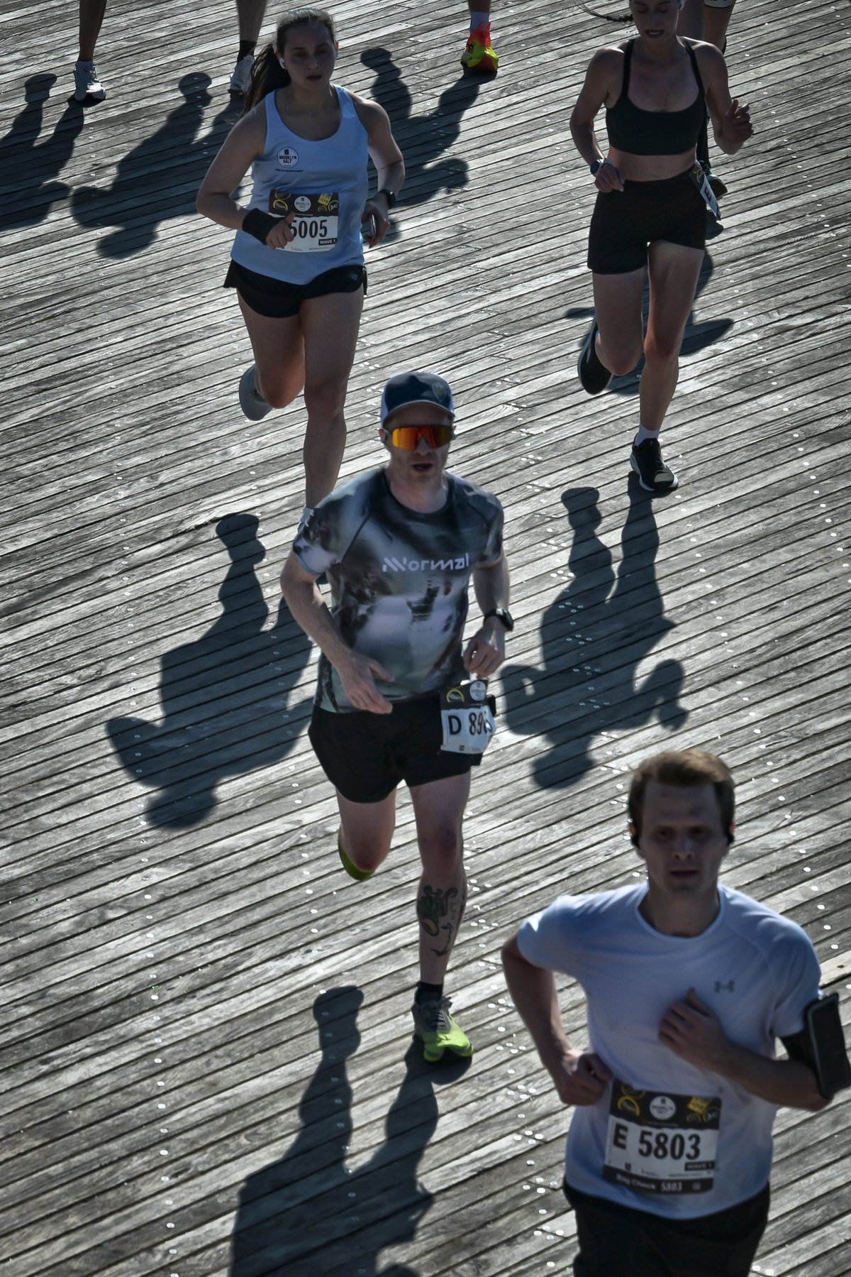 Overhead shot of runners on the Coney Island boardwalk, long shadows cast diagonally across the wooden slats, runner in grey Nnormal shirt and cap visible mid-frame