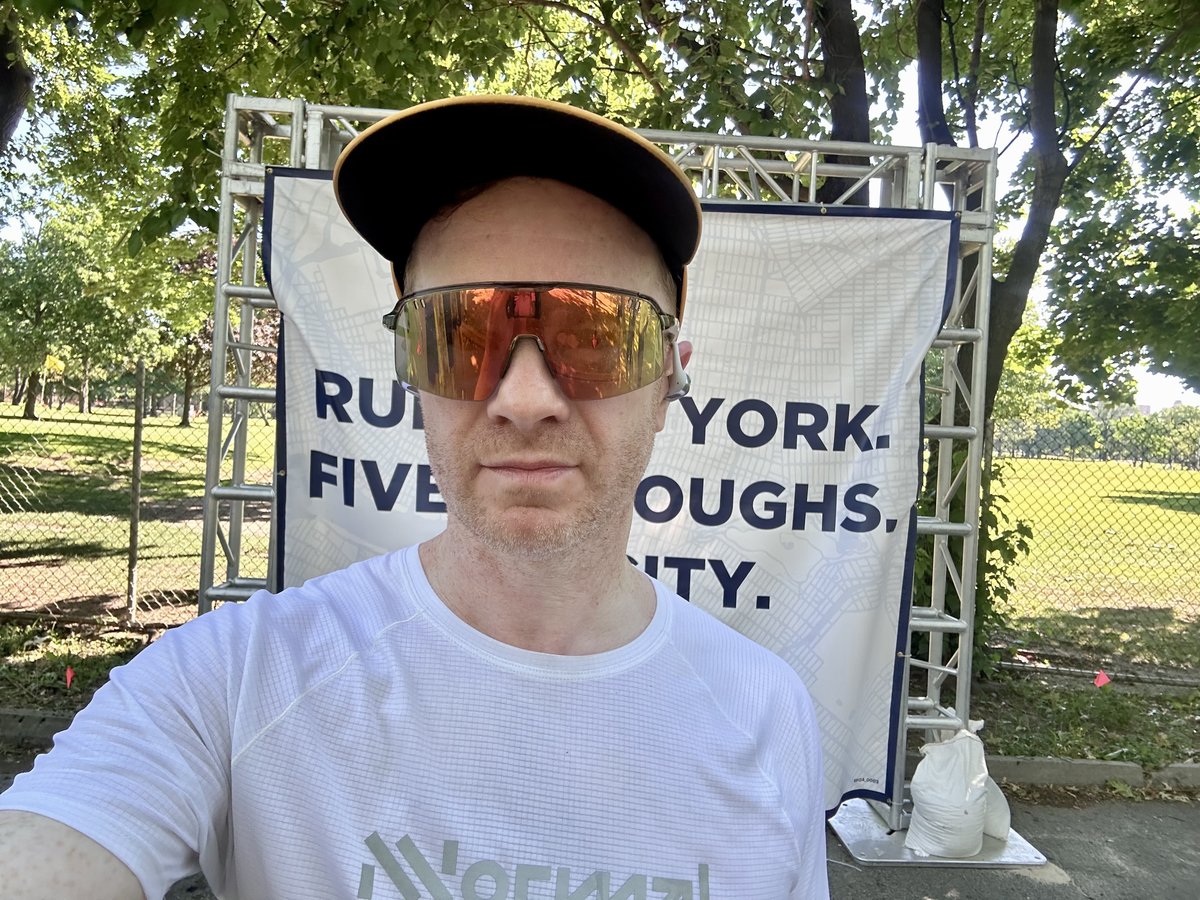 Post-race selfie in front of the Run New York Five Boroughs One City banner, orange-mirrored sunglasses and a white Nnormal shirt