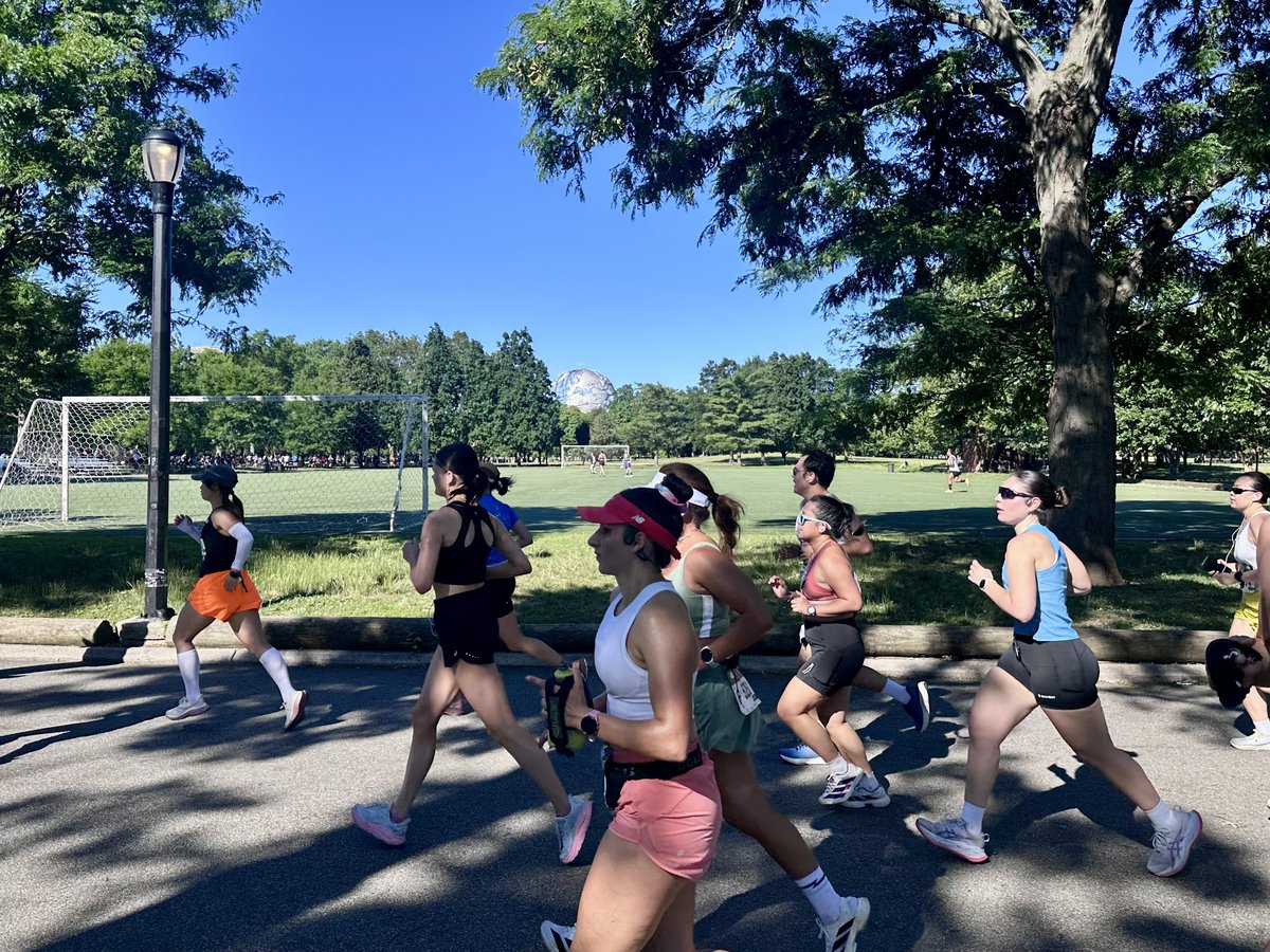 A tight pack of runners moving through a shaded section of Flushing Meadows, the Unisphere visible across an open field in the background