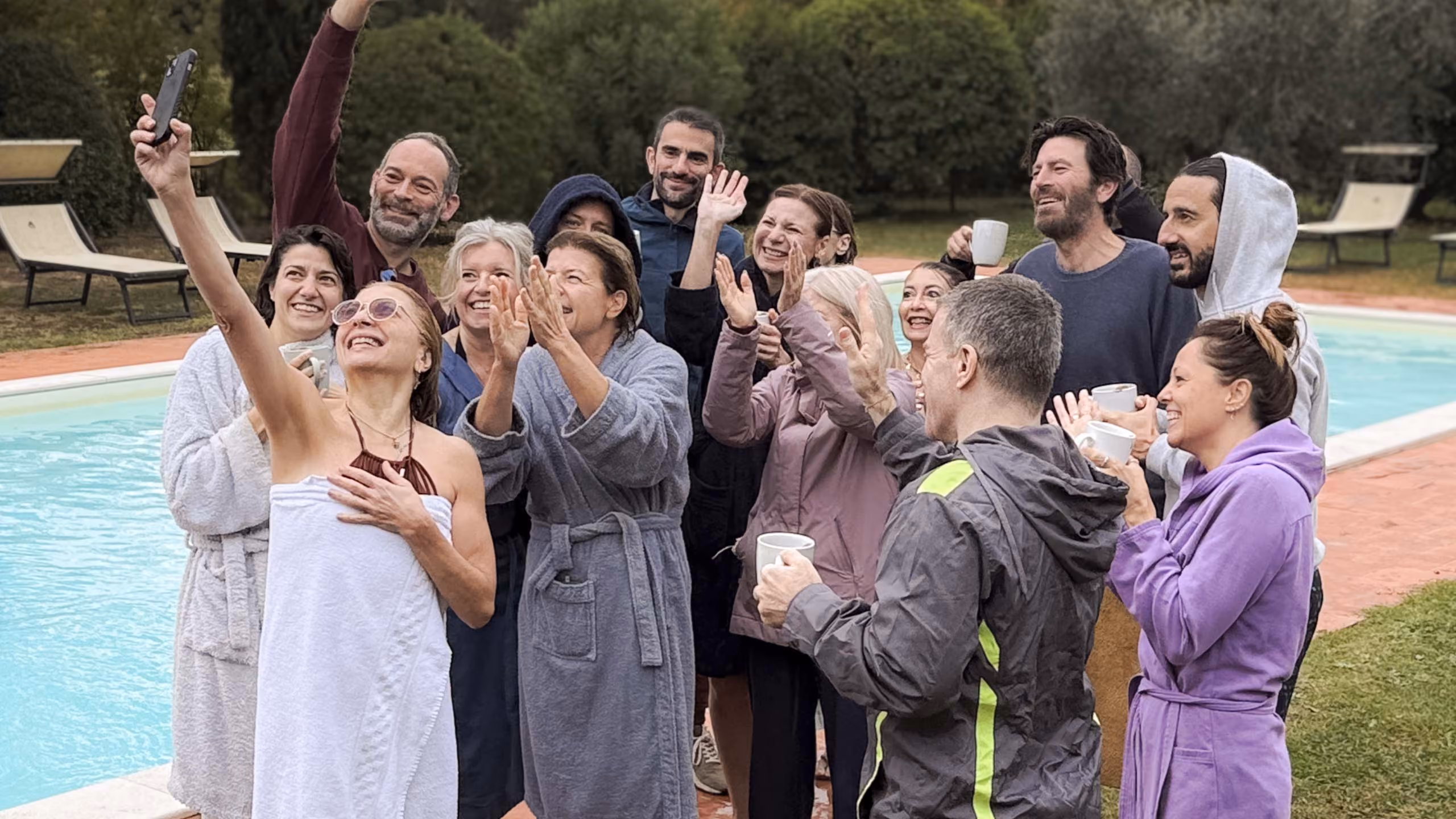 Un gruppo di persone sorridenti in accappatoi e giacche che si radunano accanto a una piscina all'aperto mentre una donna con occhiali da sole e un asciugamano bianco scatta un selfie con il telefono.