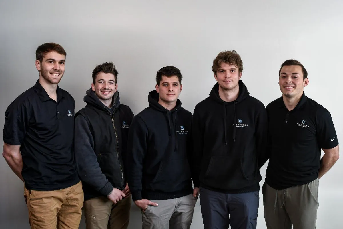 Five young men standing side by side against a plain wall, wearing black hoodies or polo shirts with a logo and smiling at the camera.