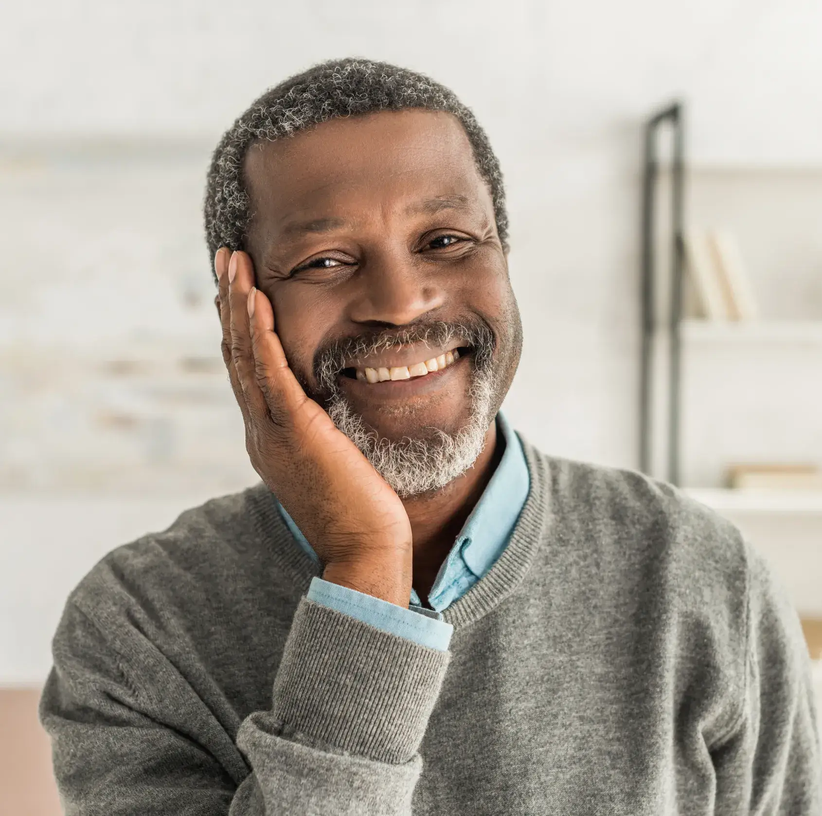 A smiling man in a gray sweater with his hand on his face.