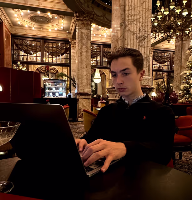 Young man typing on a laptop in a lavishly decorated room with marble pillars and chandeliers.