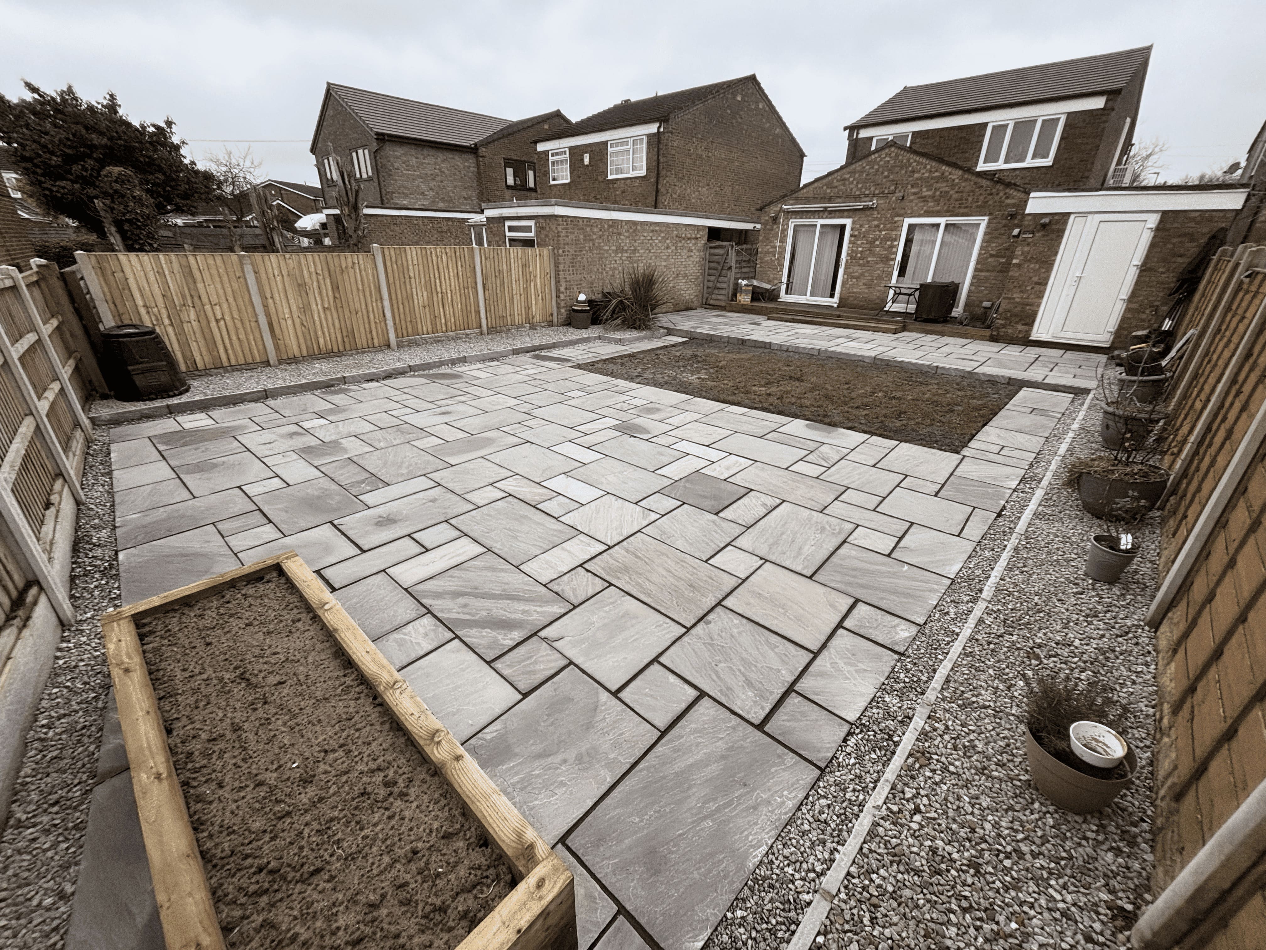 Backyard with new stone patio, gravel borders, small grass area, and multiple potted plants along fence.