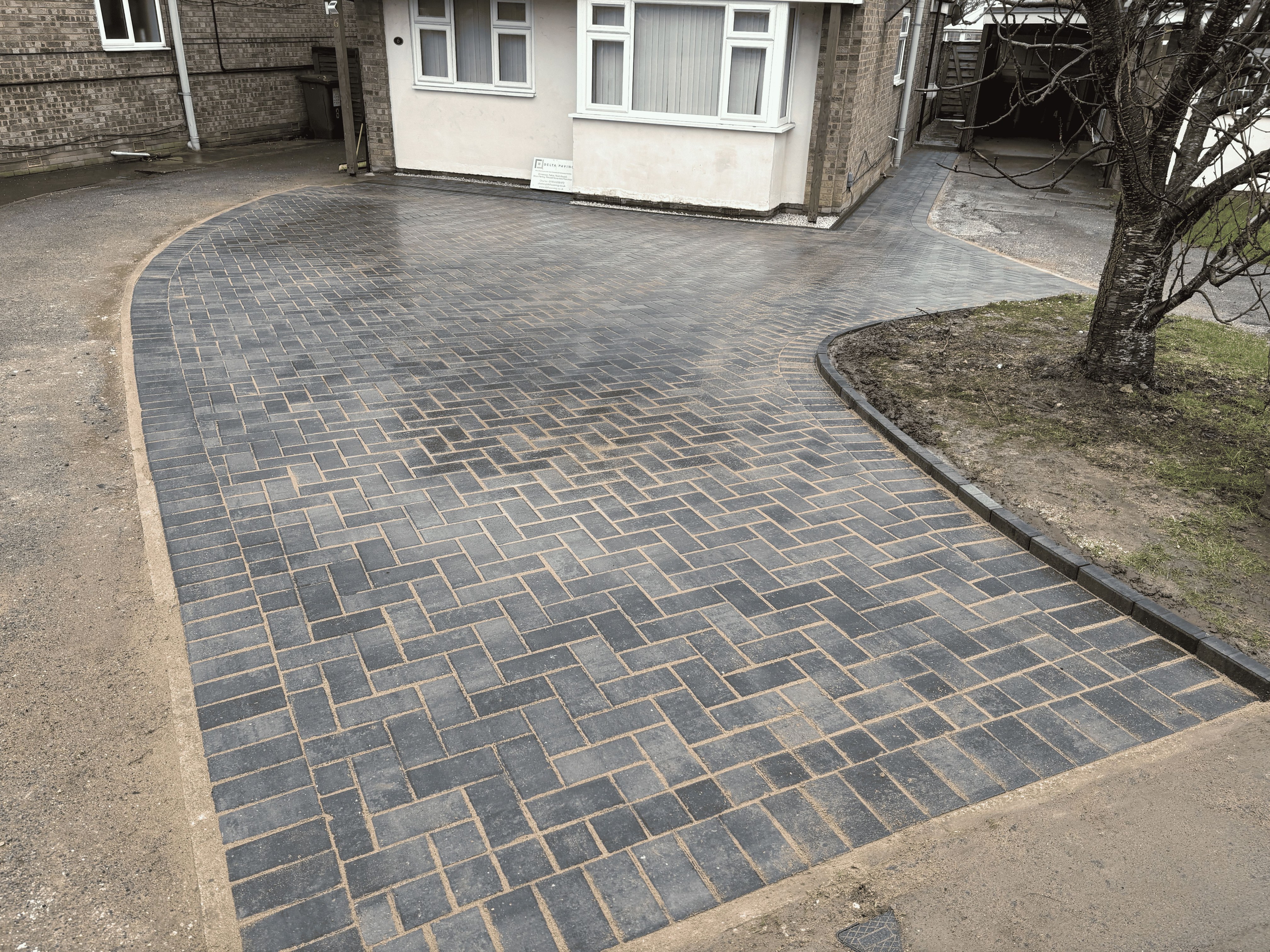 Newly paved driveway with dark bricks in a herringbone pattern next to a small garden and house.