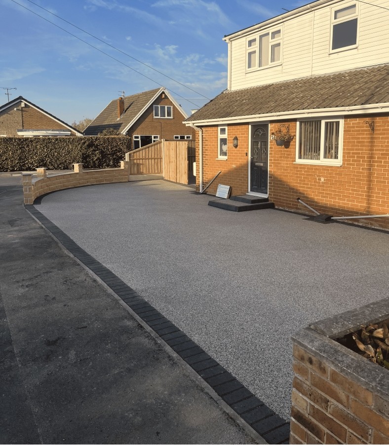 Newly resurfaced driveway with light grey resin bound gravel bordered by dark bricks outside a brick house.