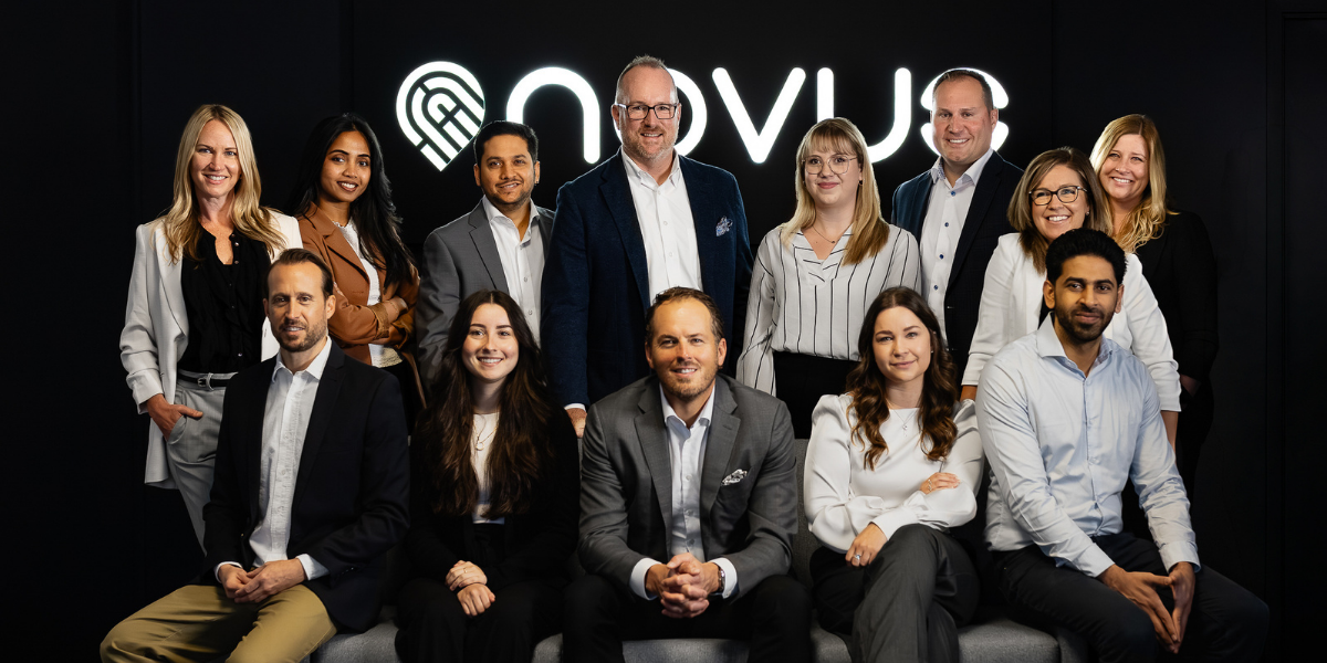 Group of diverse business professionals smiling and interacting in front of a dark wall with illuminated Novus logo.