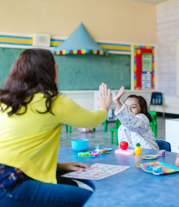 A woman and a young girl share an enthusiastic high-five while sitting in a classroom, celebrating a moment of success