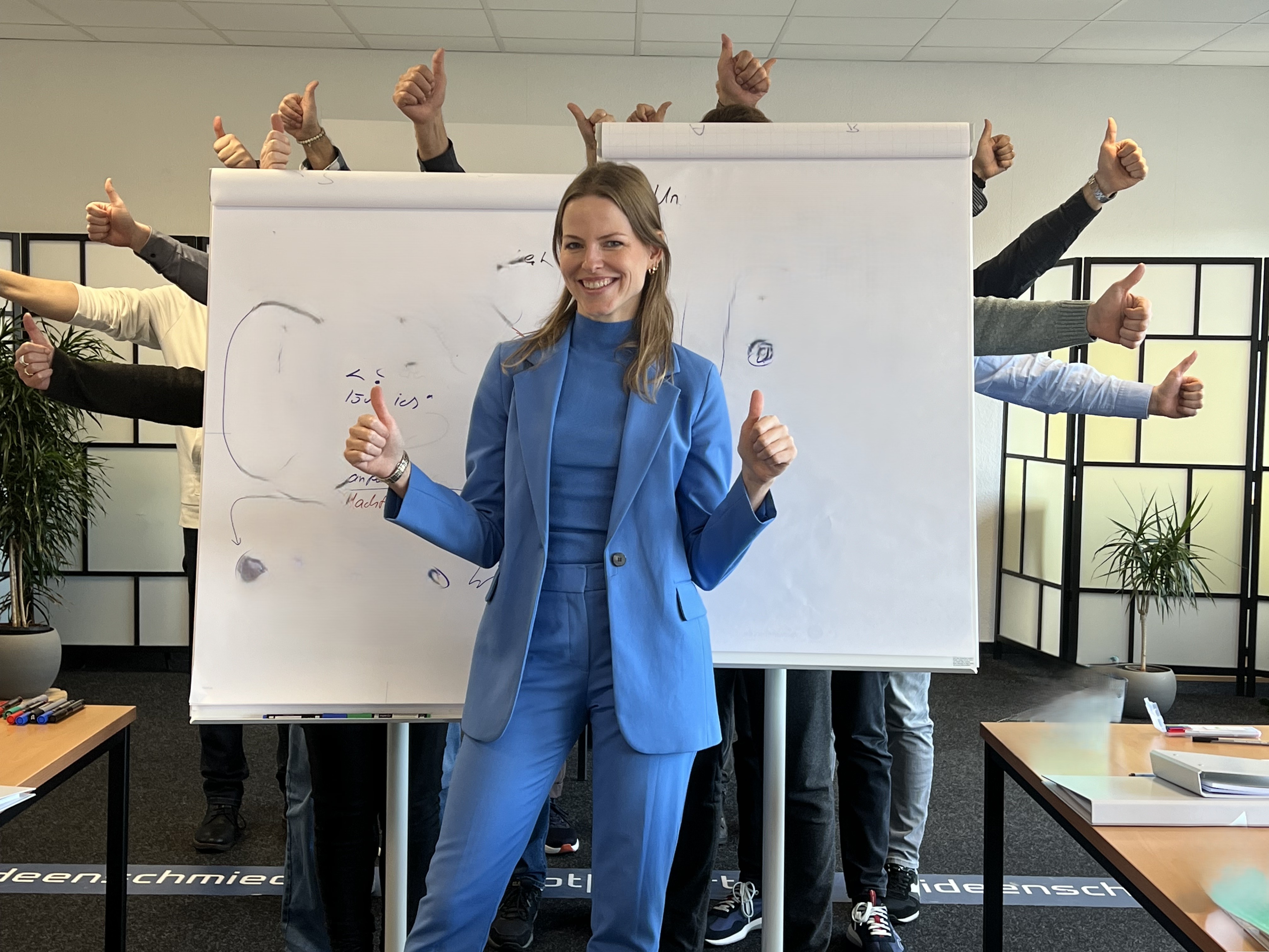 Smiling woman in blue suit standing in front of two flip charts, surrounded by people behind them giving thumbs-up gestures.