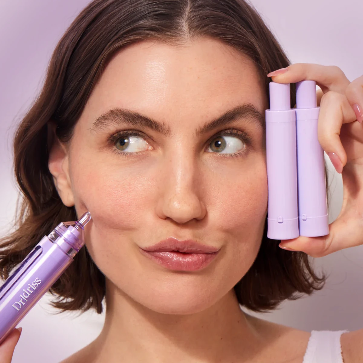Woman with short brown hair applying a lavender-colored beauty device labeled "Dr. Kiss" to her cheek, while holding two matching cosmetic tubes near her face and making a playful expression.