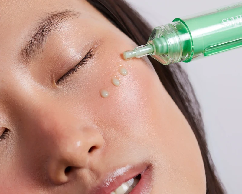 Close-up of hands dispensing Dr. Idriss "The Depuffer" Roll-On Serum onto a fingertip, against a purple background.