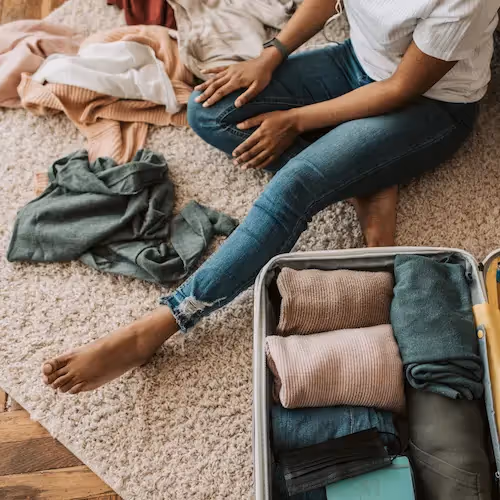 A woman sitting on a carpet packing a suitcase with neatly folded clothes while other clothing items are scattered around her.