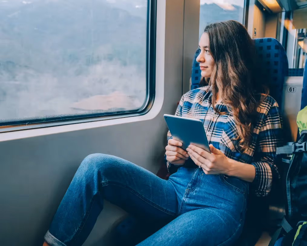 A woman sitting by a train window holding a tablet, gazing at the scenery outside.
