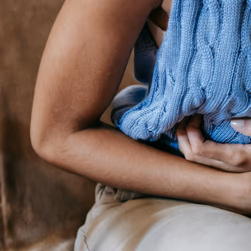Woman in blue cable-knit sweater sits on beige blanket, holding stomach, suggesting IBS discomfort. Neutral brown background, cozy setting.