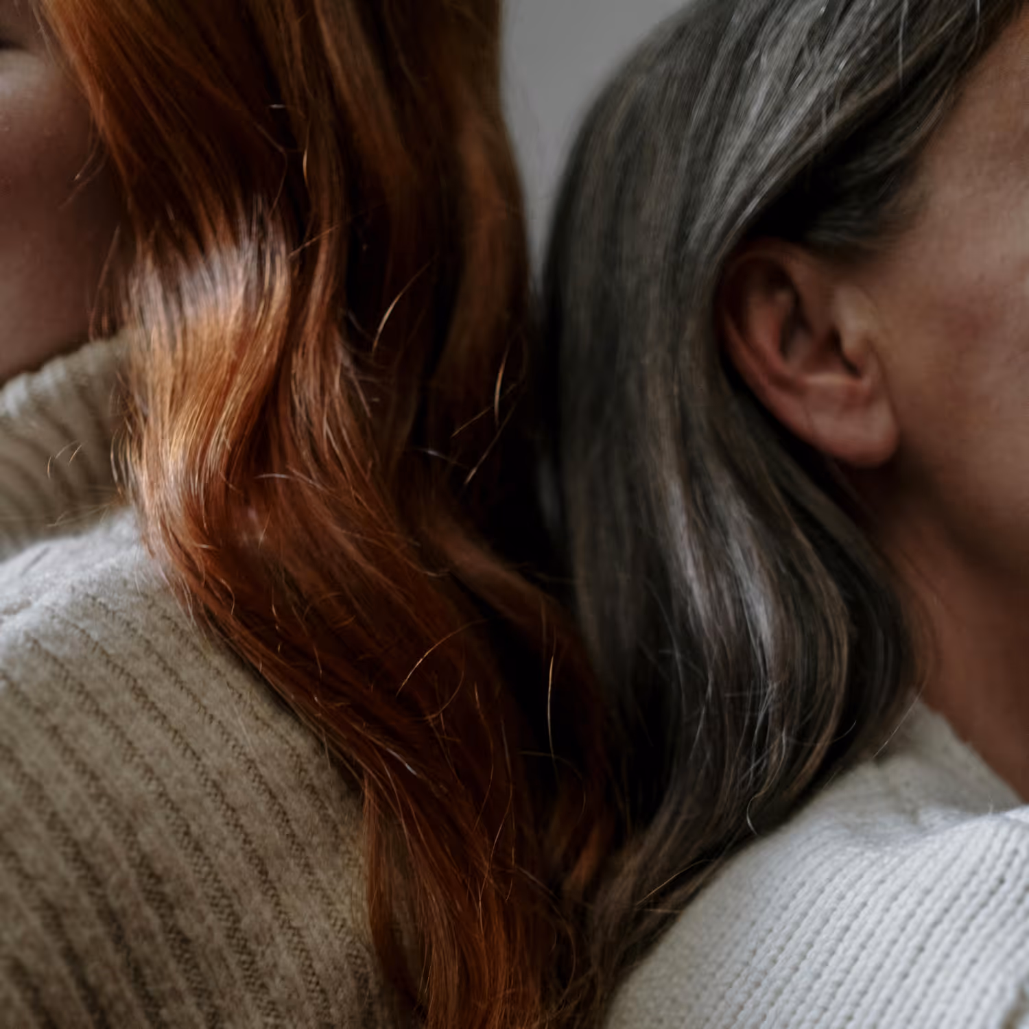 Two women sitting back to back, one with red hair and the other with gray hair, both wearing light knit sweaters.