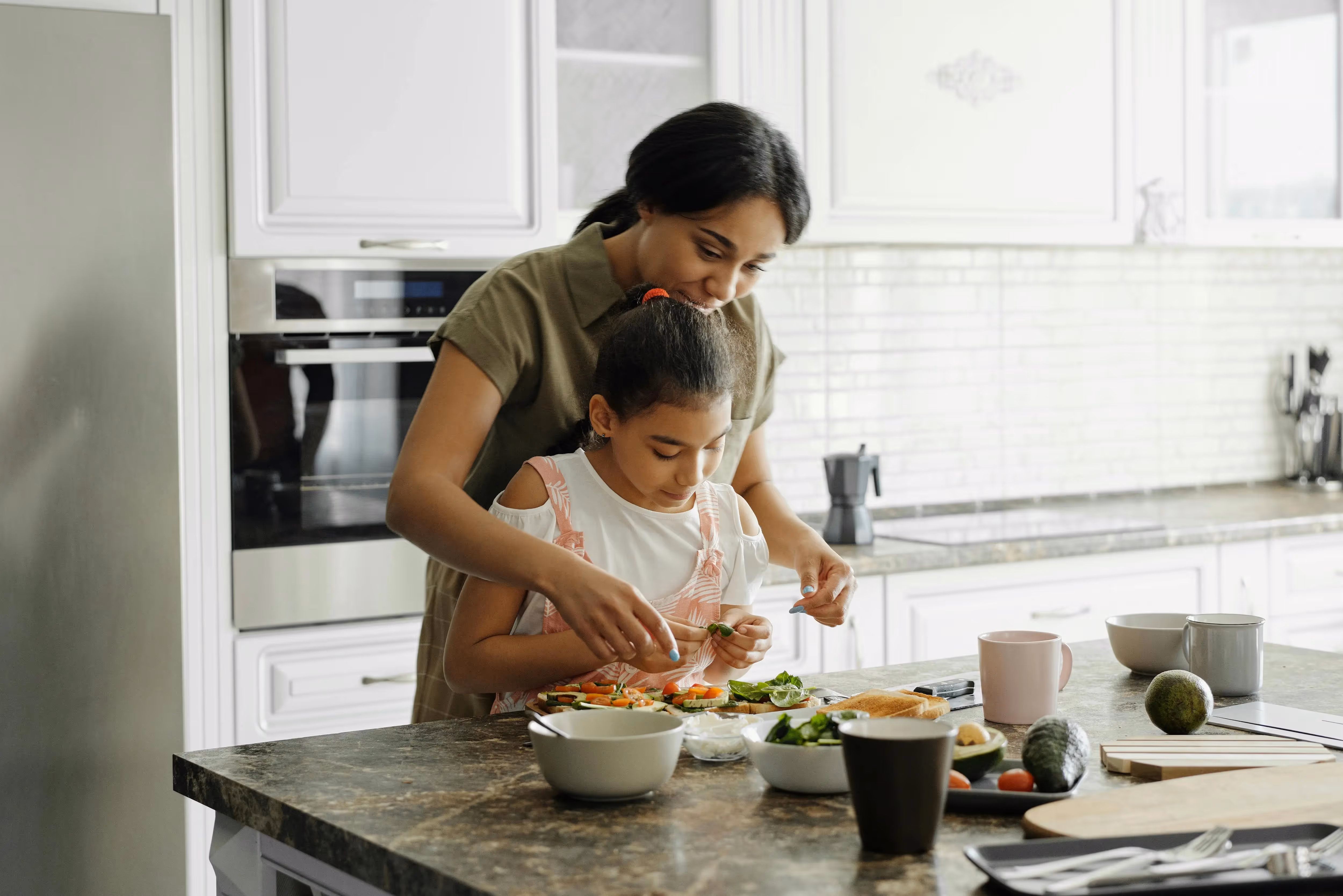 A woman and young girl prepare a healthy meal together in a modern kitchen, arranging vegetables on bread at a counter surrounded by bowls of fresh ingredients.