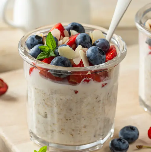 A glass jar of gut-friendly overnight oats layered with yogurt, fresh blueberries, sliced strawberries, almonds, and a sprig of mint, with a spoon inside.