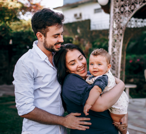 A happy couple embraces their baby in an outdoor garden, smiling warmly as they enjoy a tender family moment together.