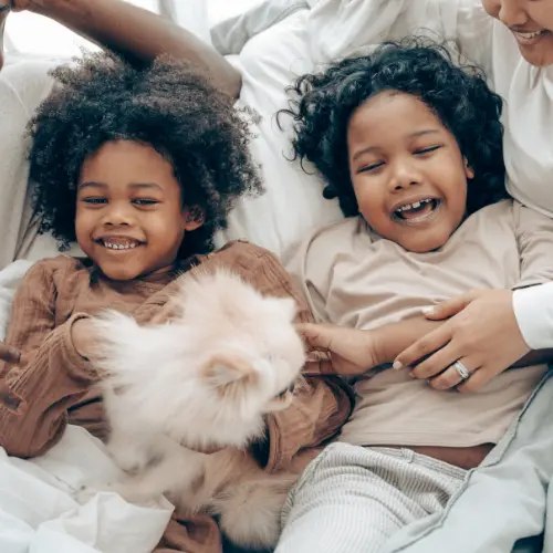 Two smiling children lie on a bed with a fluffy white dog, cuddled close to a parent’s arm.