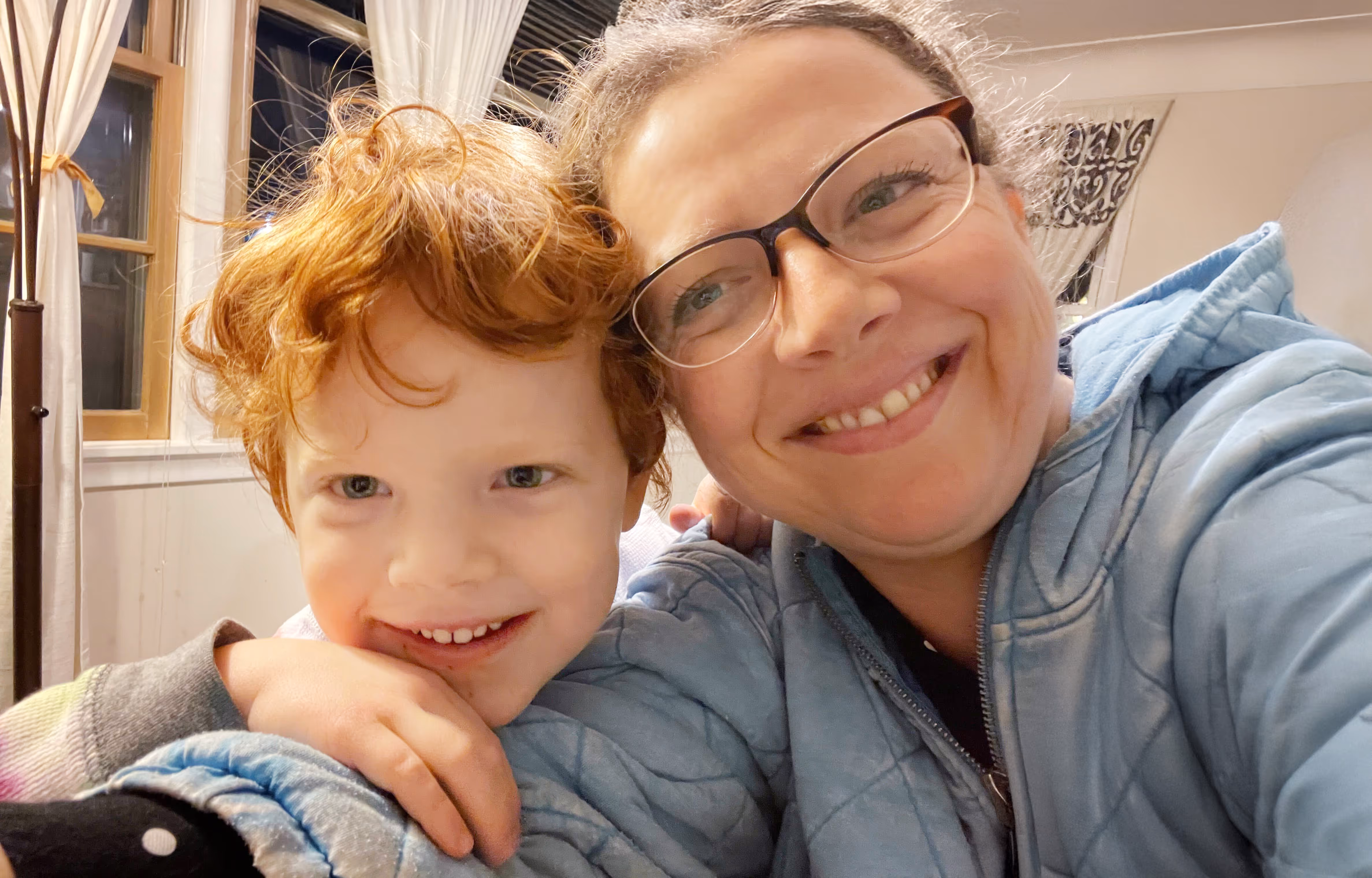 Woman with glasses and child with curly red hair smiling together indoors in a selfie.