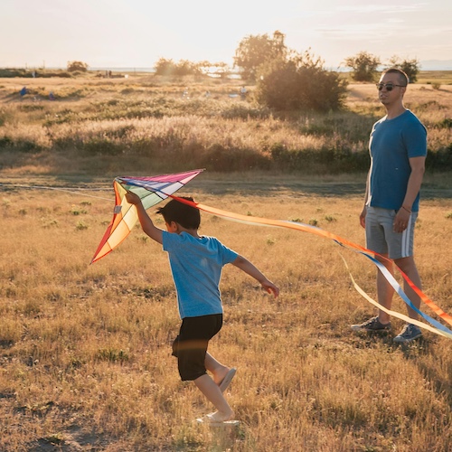 Child running with colorful kite in golden field at sunset, adult watches nearby amid dry grassland and distant trees.