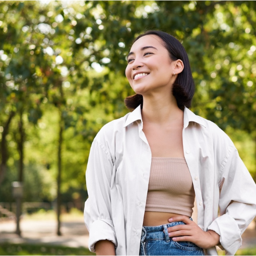 Woman smiling outdoors in white shirt, beige top, and jeans with trees in background.